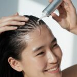 Scalp Detox A smiling person applies serum or oil to their scalp with a dropper, massaging it into wet hair.