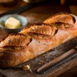 French Bread Recipe A Freshly Baked Loaf Of French Bread Sits On A Wooden Cutting Board, With A Butter Knife Placed Beside It And A Small Bowl Of Butter In The Background. The Bread Has A Golden Crust With Three Diagonal Slashes Along Its Top.