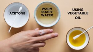 Hand With Adhesive Stuck On The Finger And Bowls Labeled Acetone, Warm Soapy Water, And Vegetable Oil For Adhesive Removal Methods.