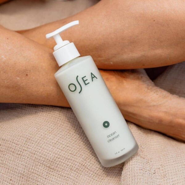 Osea Ocean Cleanser Bottle Lying On A Textured Beige Cloth With A Person’s Legs In The Background.