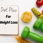 An Open Notebook With &Quot;The Menopause Diet 5 Day Plan For Weight Loss&Quot; Written On It, Surrounded By Healthy Items Like Tomatoes, A Tape Measure, Green Dumbbells, And Uncooked Pasta On A White Surface.