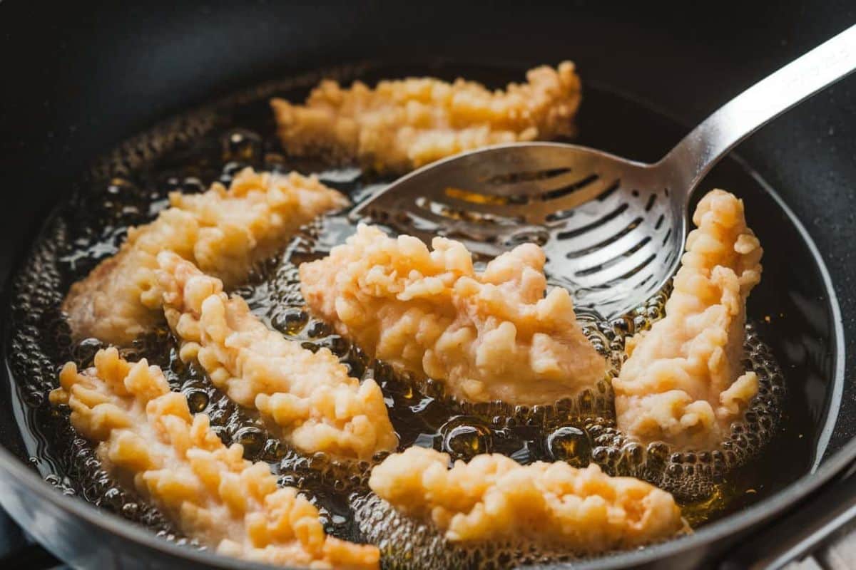 Chicken tempura frying in hot oil with a slotted spoon used for turning and removing pieces.