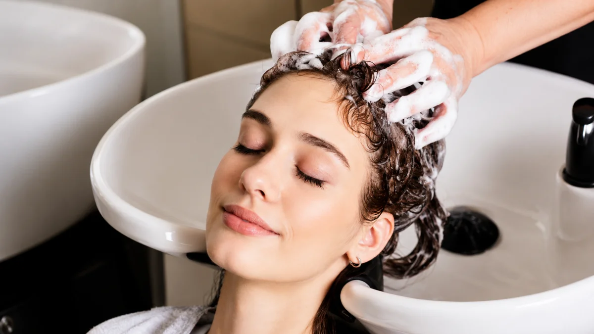 A Relaxed Young Woman With Curly Dark Hair Leaning Back At A Salon Wash Basin While A Stylist Performs A Foamy Scalp Detox Shampoo Massage, With Eyes Closed And A Peaceful Expression.