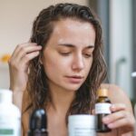 Woman examining a bottle of hair oil for dry scalp, surrounded by various skincare and haircare products on a bathroom counter.