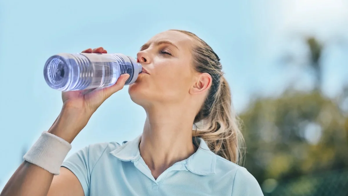 A Woman In A Light Blue Polo Shirt And Wrist Sweatband Drinking Water From A Clear Plastic Bottle Outdoors In The Heat, Representing The Importance Of Hydration In A Daily Body Care Routine.