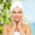 A Step-by-Step Body Care Routine for Glowing and Healthy Skin 7 A Smiling Woman With A White Towel Wrapped Around Her Head Gently Touching Her Glowing, Healthy Face After Completing Her Daily Body Care Routine, With A Fresh Green And Blue Outdoor Background.