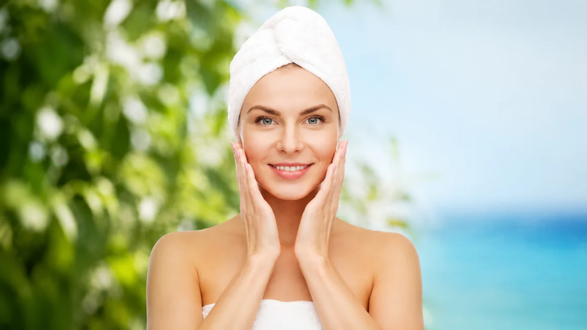 A Smiling Woman With A White Towel Wrapped Around Her Head Gently Touching Her Glowing, Healthy Face After Completing Her Daily Body Care Routine, With A Fresh Green And Blue Outdoor Background.