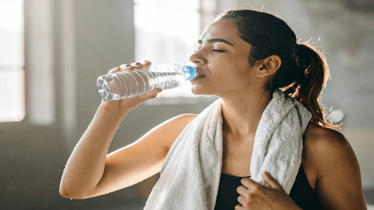 How to Revive Dull Skin: A Step-by-Step Guide to Restoring Your Natural Glow 3 A Young South Asian Woman With A White Gym Towel Around Her Neck Drinking From A Plastic Water Bottle After A Workout In A Bright Gym Setting — Illustrating How Proper Daily Hydration Is One Of The Most Effective And Natural Ways To Combat Dull Skin From The Inside Out.