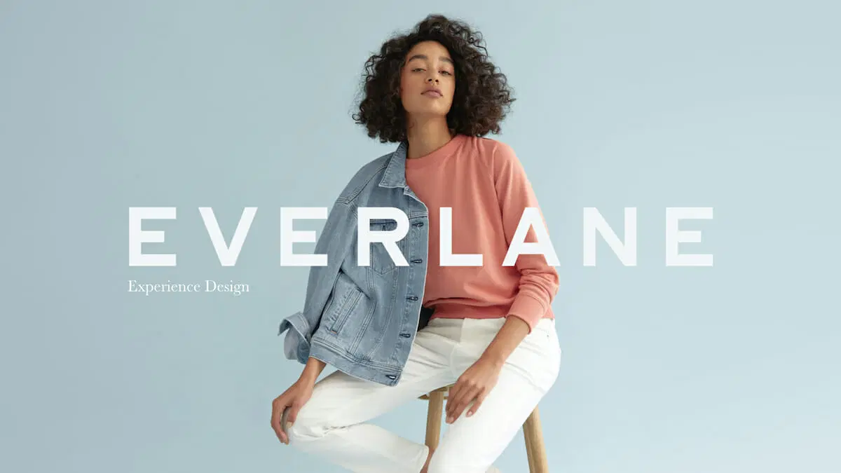 A Woman With Natural Curly Hair Sitting On A Wooden Stool Wearing An Everlane Denim Jacket Over A Coral Pink Sweatshirt With White Jeans, Posed Confidently Against A Soft Light Blue Background In An Everlane &Quot;Experience Design&Quot; Brand Campaign Image.