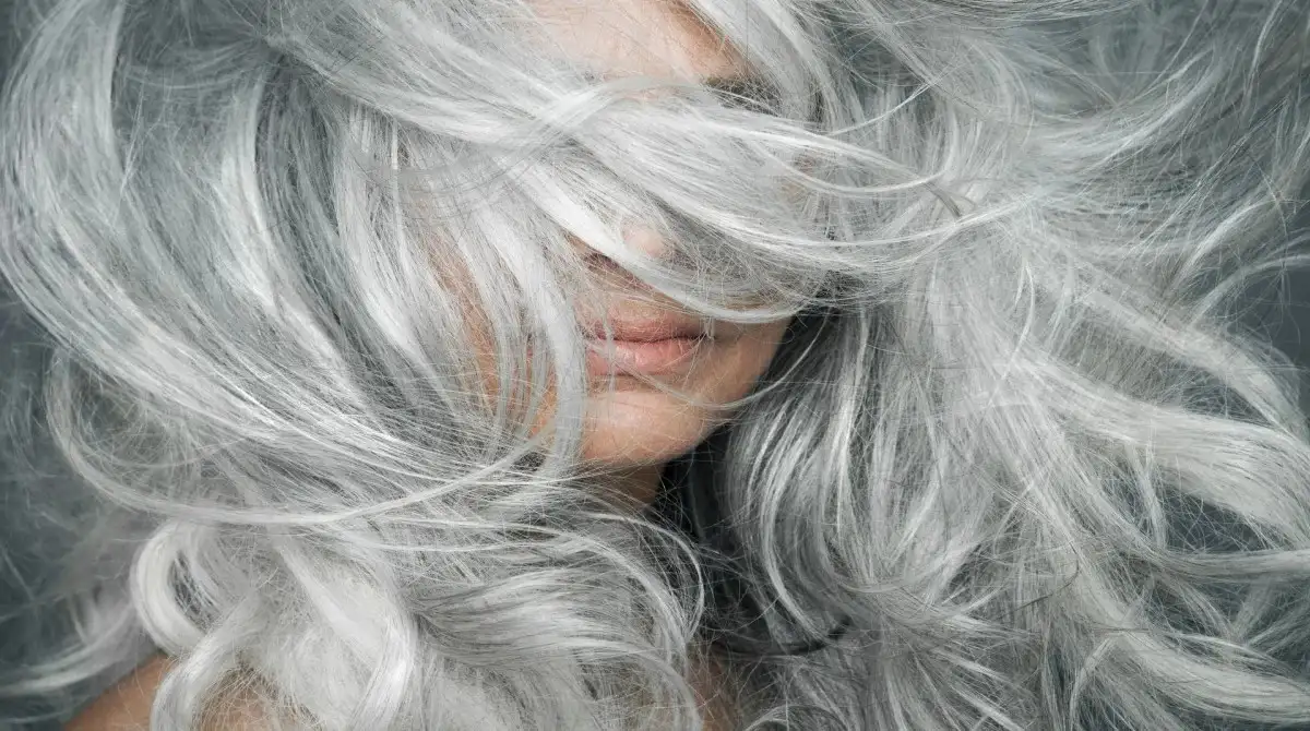 Grey Hair Blending Is Becoming the Go-To Beauty Trend for a Softer, Younger Look 2 A Close-Up Portrait Of A Woman Smiling Outdoors As Natural Strands Of Grey Hair Softly Blow Across Her Face, Showcasing A Vibrant Silver Texture.