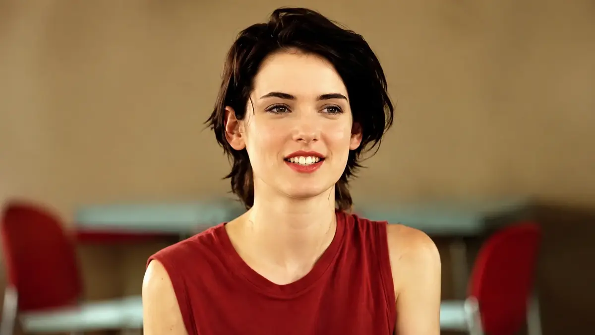 Winona Ryder Smiling In A Sleeveless Red Top In A High-Definition 16:9 Portrait With A Soft Blurred Indoor Background.